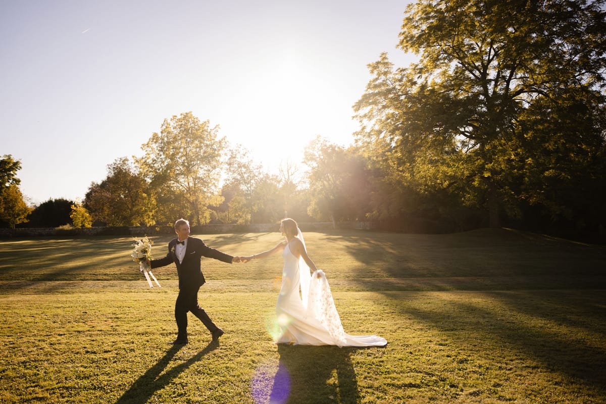 Couple walking hand in hand across estate lawn at golden hour, groom in black tuxedo and bride in fitted ivory gown with t...