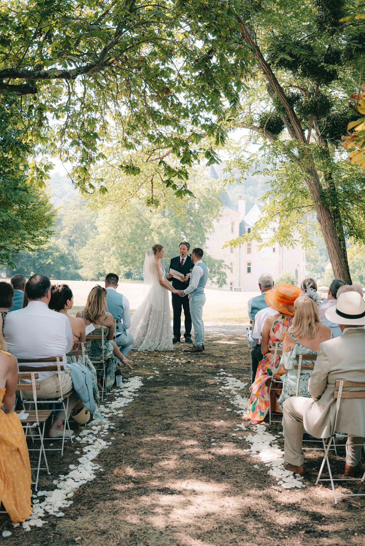 Outdoor wedding ceremony on chateau grounds with bride in lace dress and cathedral veil facing groom under mature trees