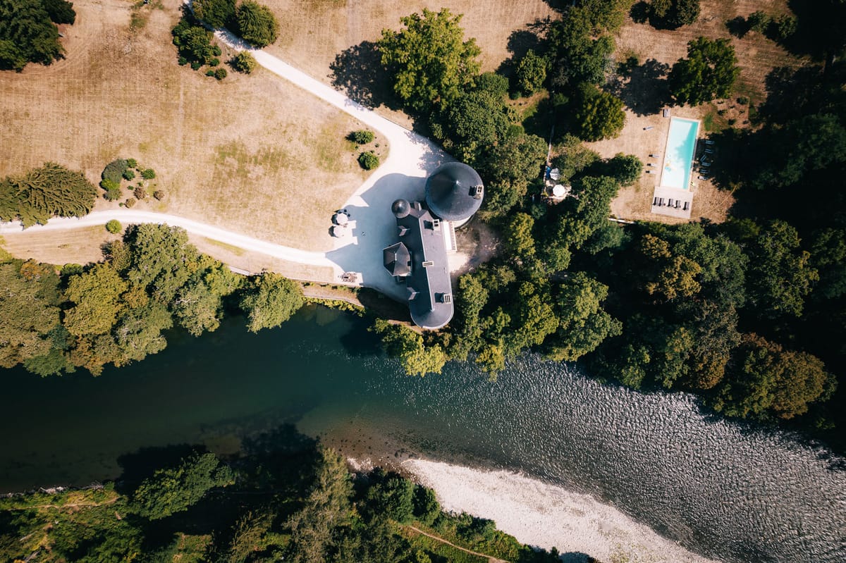 Aerial drone view of riverside French estate with round tower, swimming pool, and gravel driveway through open grounds