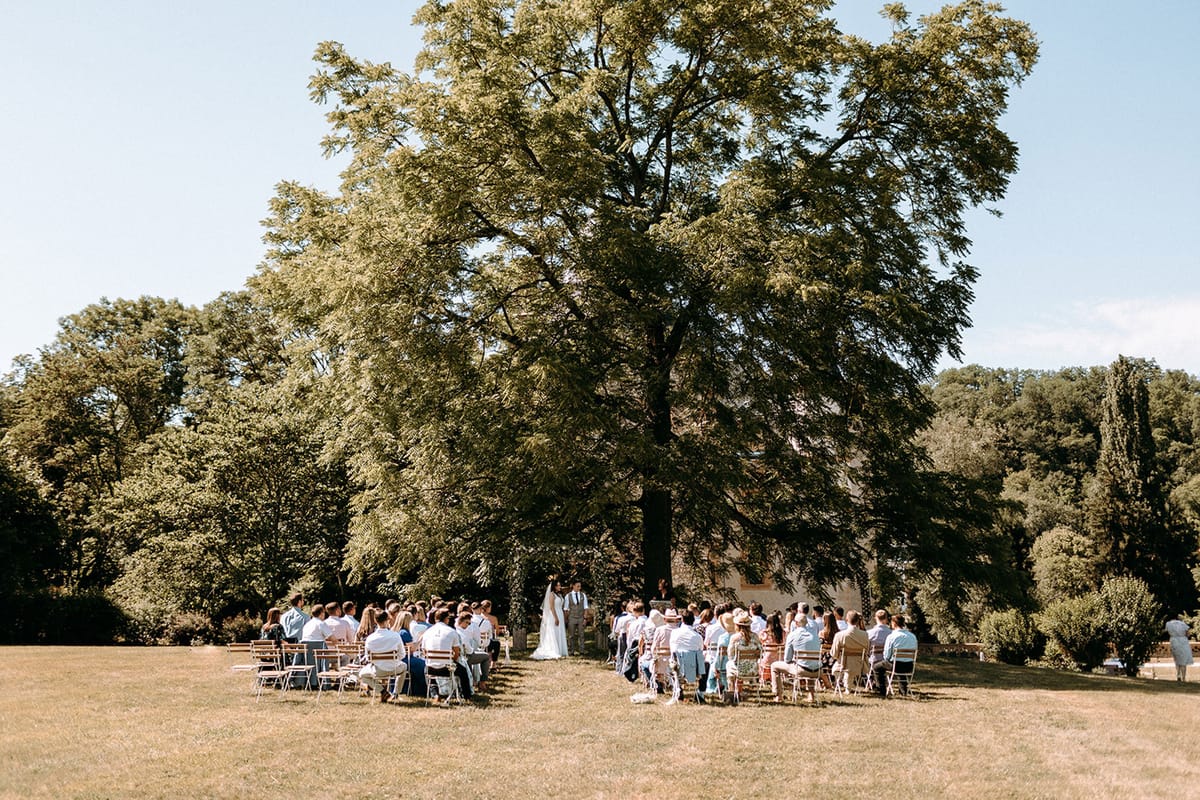 Outdoor wedding ceremony under a large tree with guests seated on wooden chairs on a chateau lawn