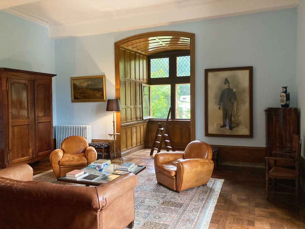 Chateau salon with pale blue walls, diamond-pane leaded window, tan leather club chairs, and antique armoire