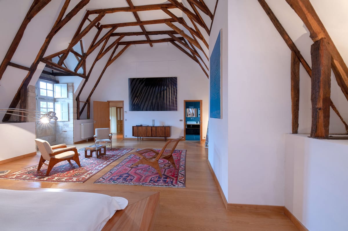Attic bedroom with exposed timber trusses, mid-century armchairs, kilim rugs, and teal bathroom doorway