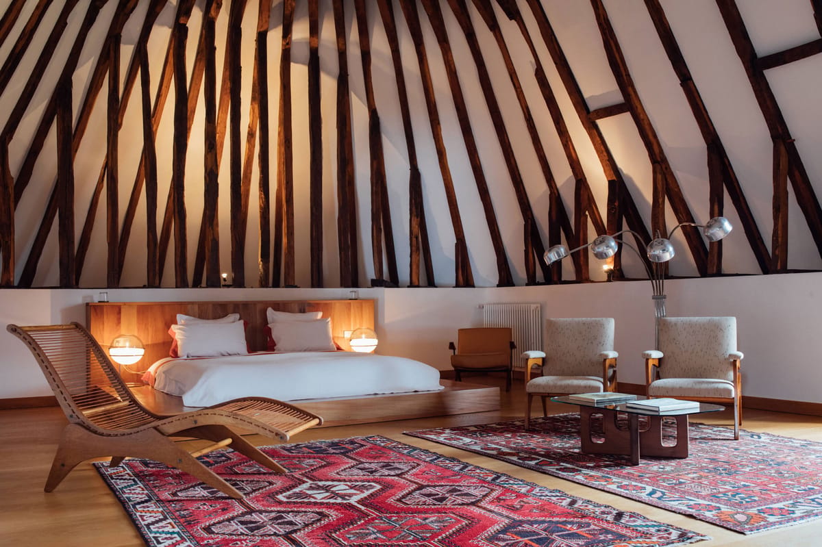 Vaulted bedroom with exposed timber trusses, mid-century rattan chaise, rust cushions, and kilim rugs