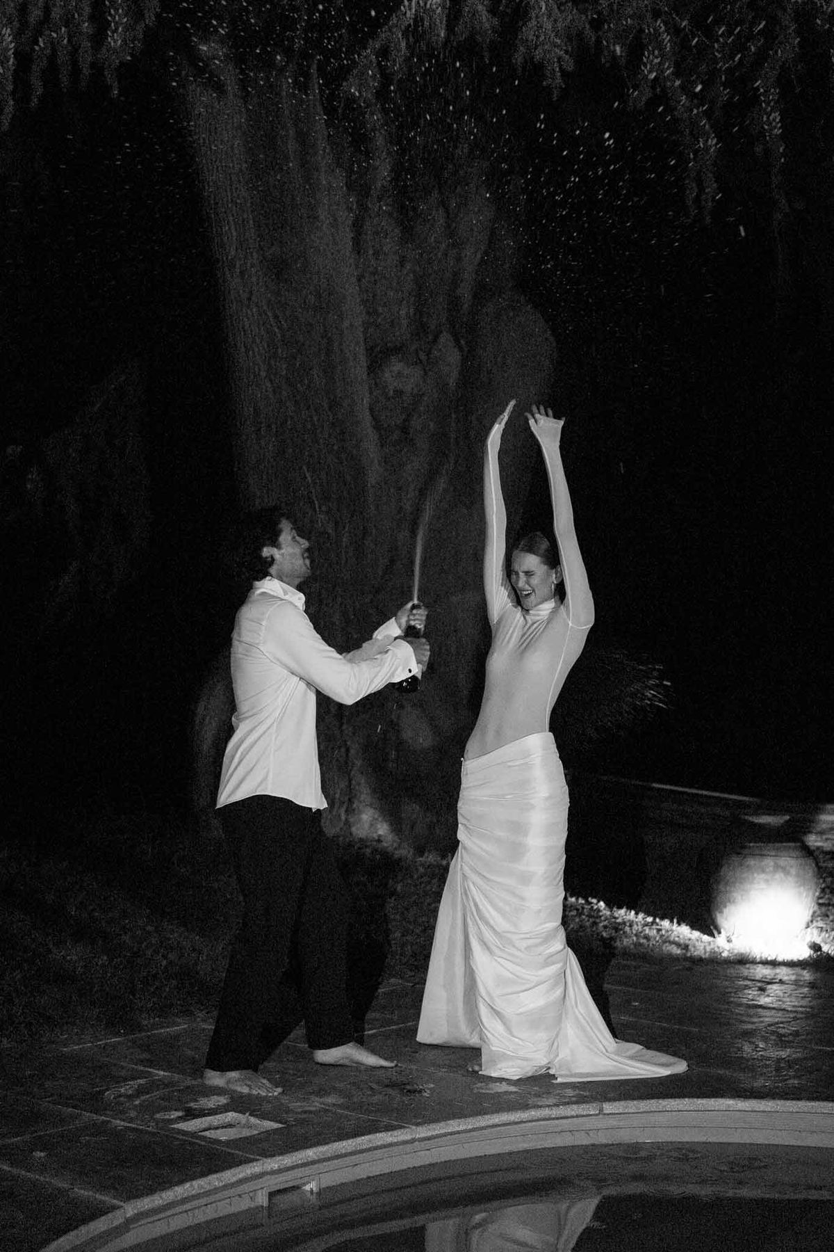 Black and white nighttime portrait of couple sabering champagne bottle by pool with dramatic spray
