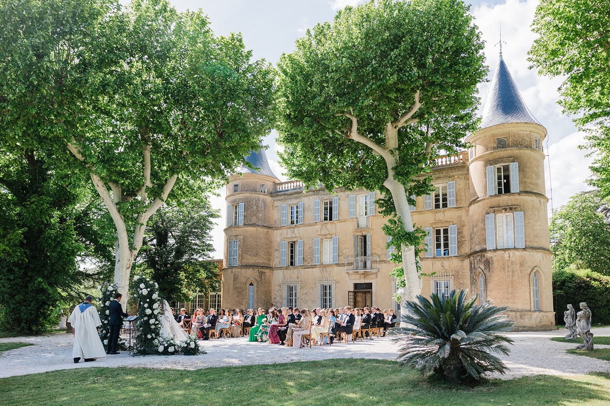 Outdoor ceremony with 60-plus guests before golden stone chateau with white floral arch and wooden chairs