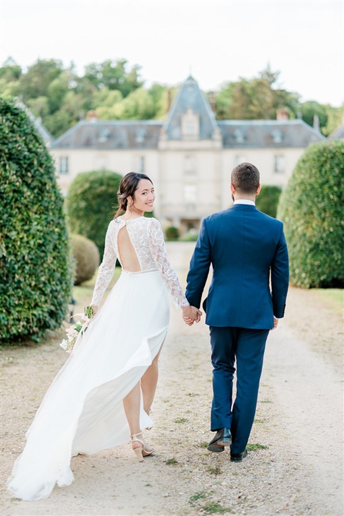 Couple walking toward chateau on topiary-hedged gravel path with bride glancing back in lace gown