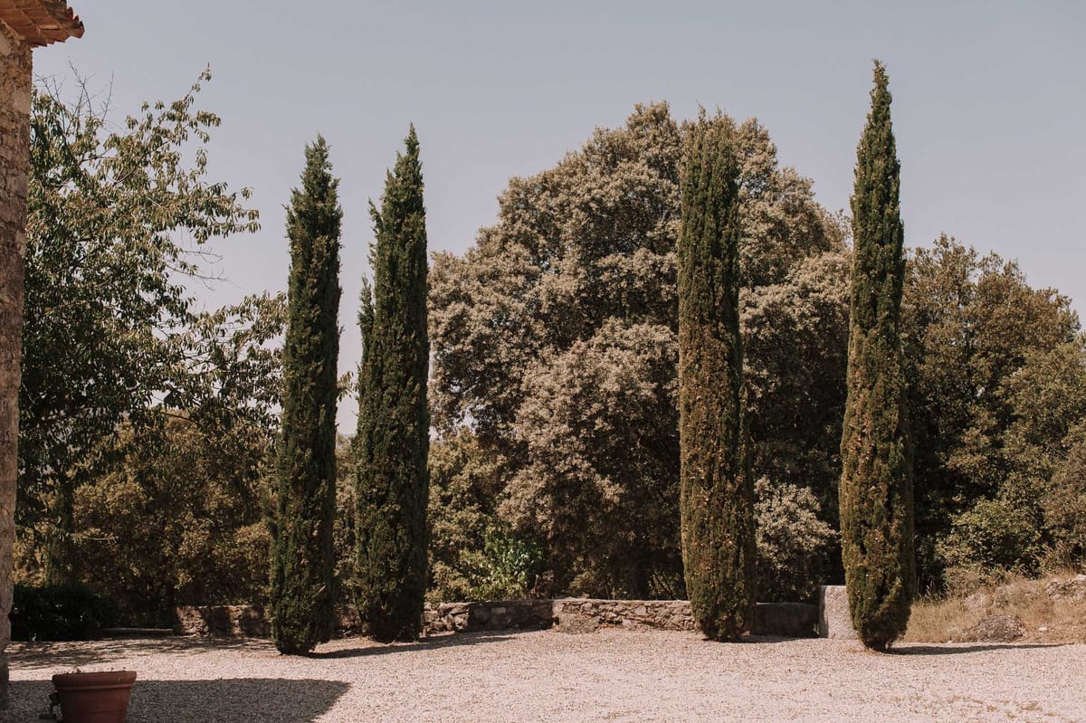 Gravel courtyard lined with Italian cypress trees beside stone building with terracotta roof and low stone wall