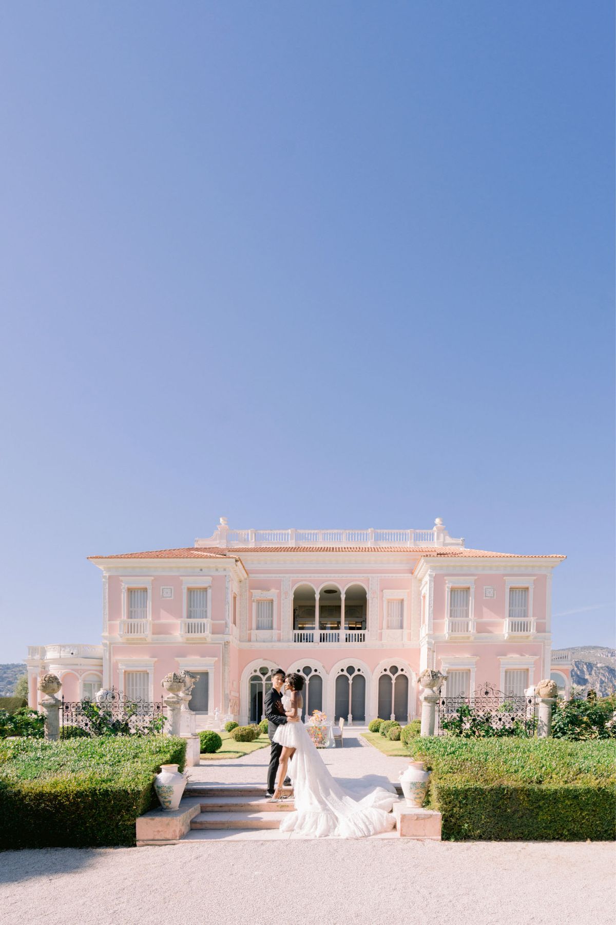 Bride in strapless ball gown and groom in black suit on garden steps of pink villa with cathedral train