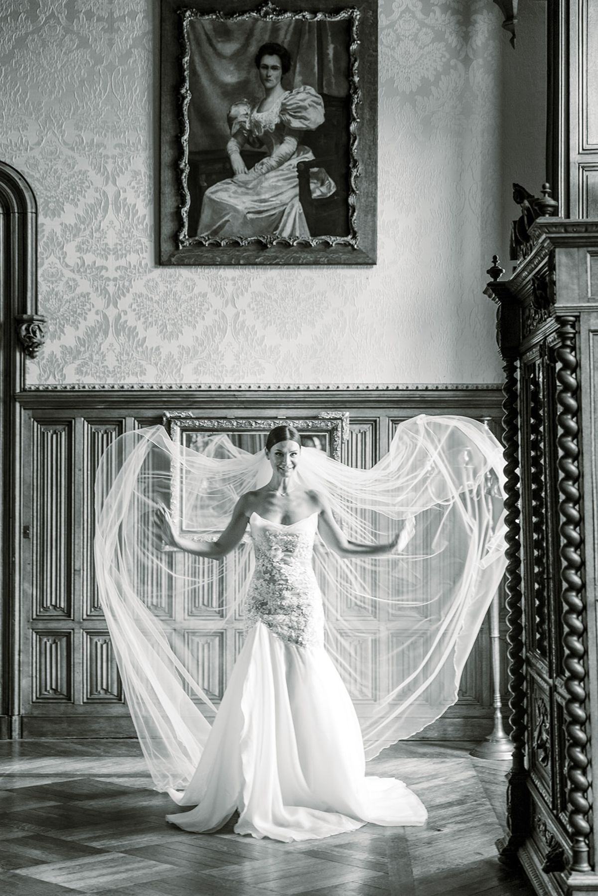 Bride lifting veil beneath portrait in panelled salon at Chateau Challain, Loire Valley