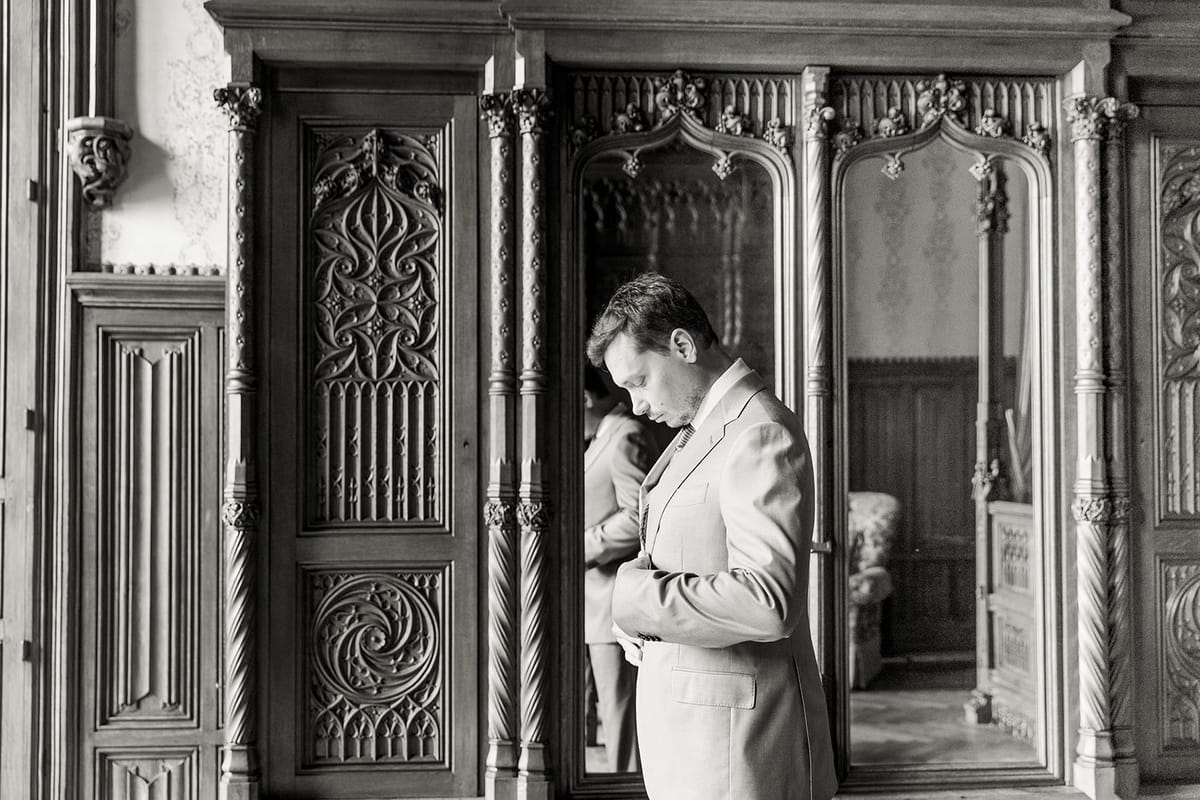 Groom buttoning suit jacket beside carved armoire at Chateau Challain, Loire Valley