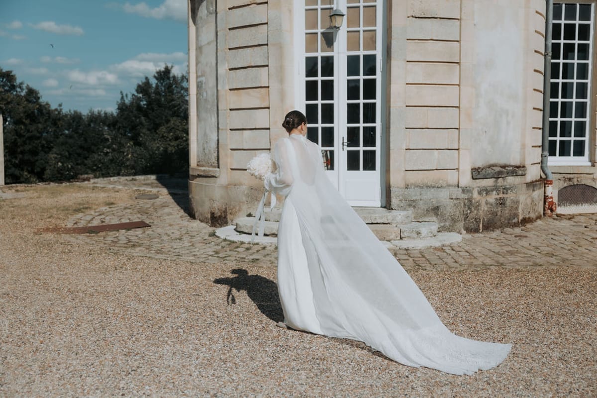 The bride walks across the gravel towards the château, her veil trailing behind her