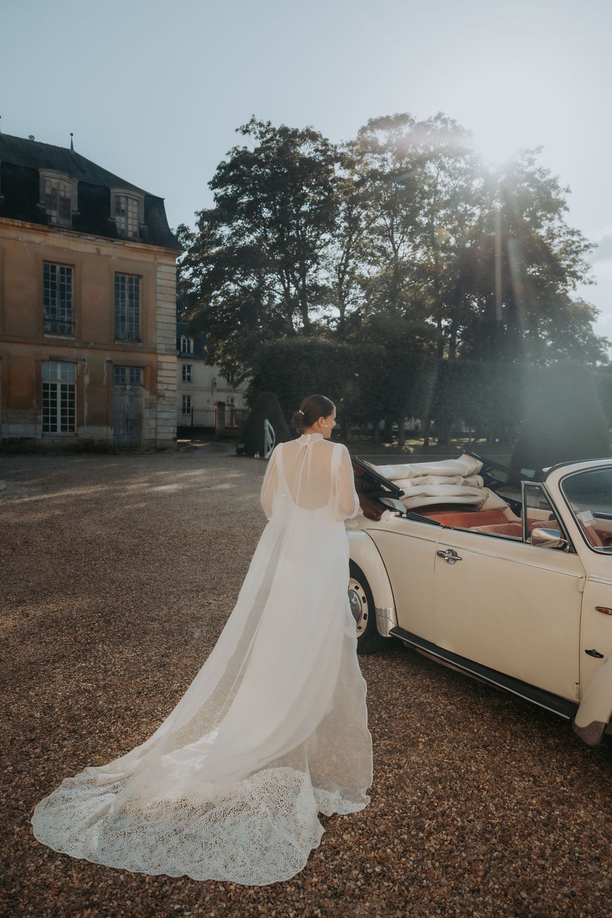 The bride beside a cream vintage car in front of the château, seen from behind