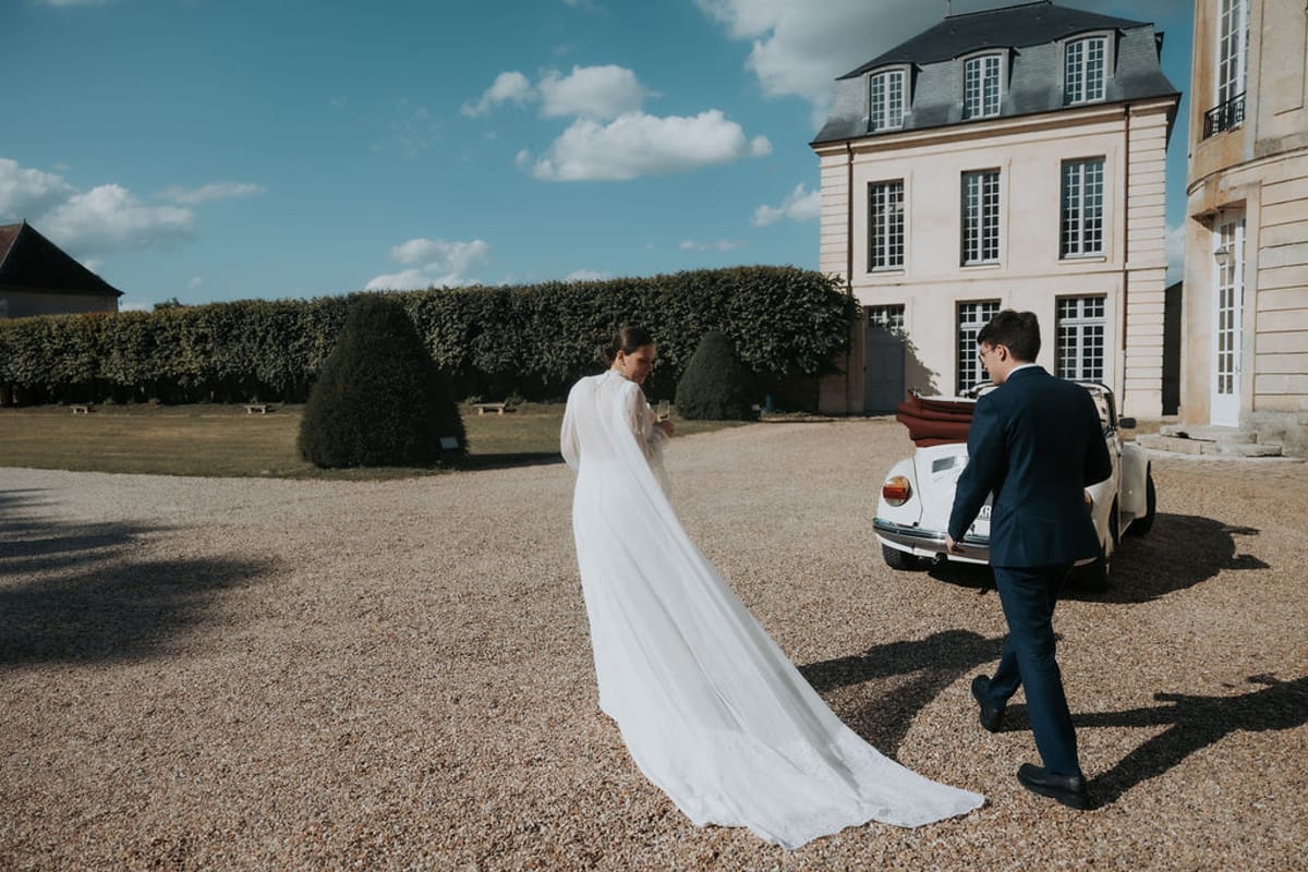 The bride and groom walk across the gravel courtyard towards the château, a vintage car beside them