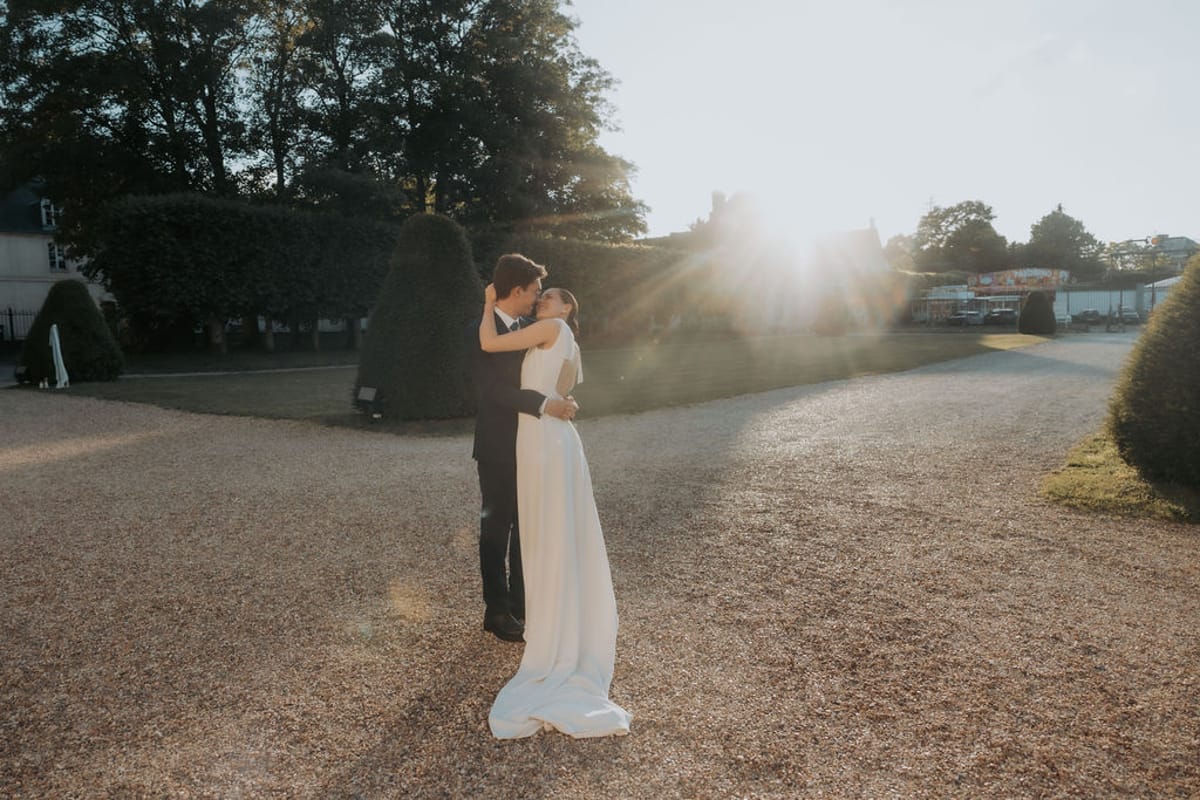 The bride and groom embrace on the gravel drive in the evening sun