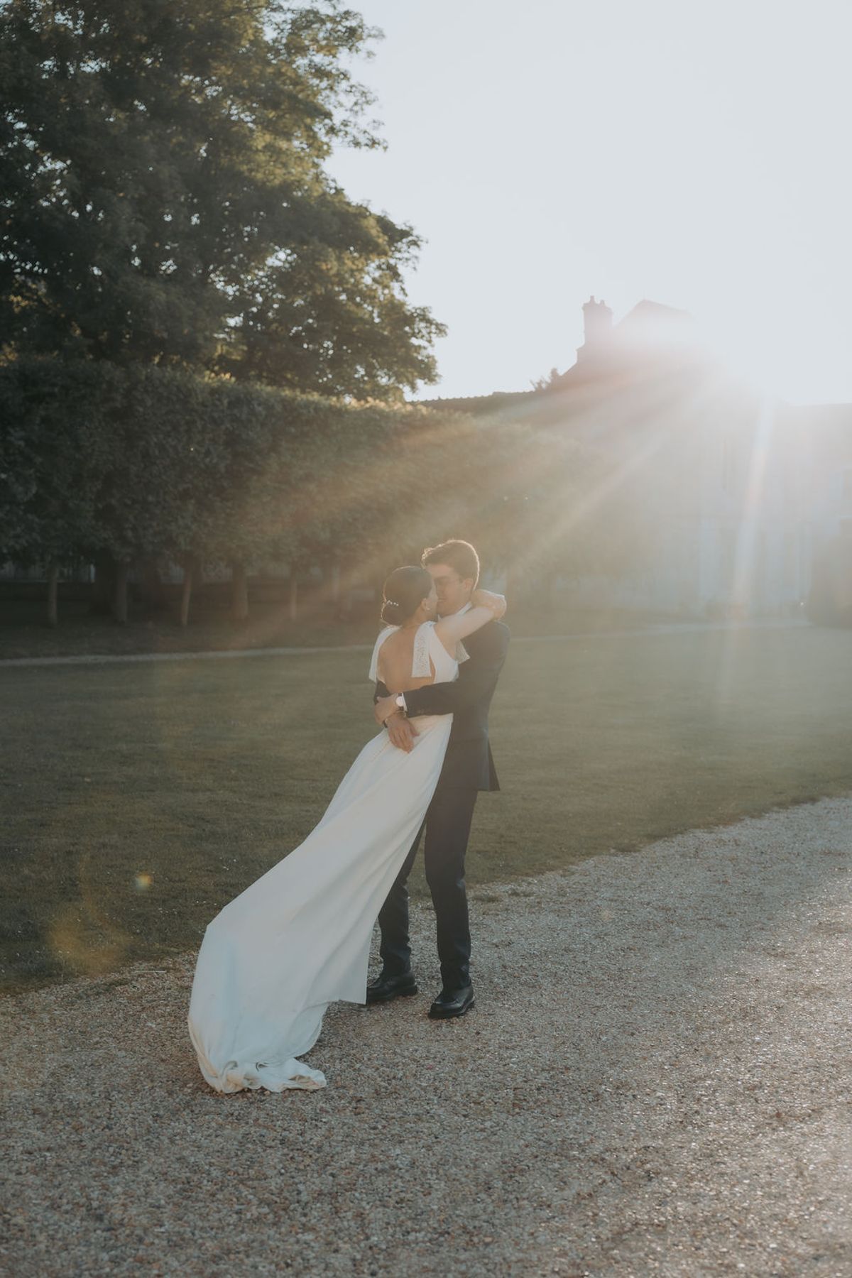The bride and groom embrace on the gravel with the low sun behind them