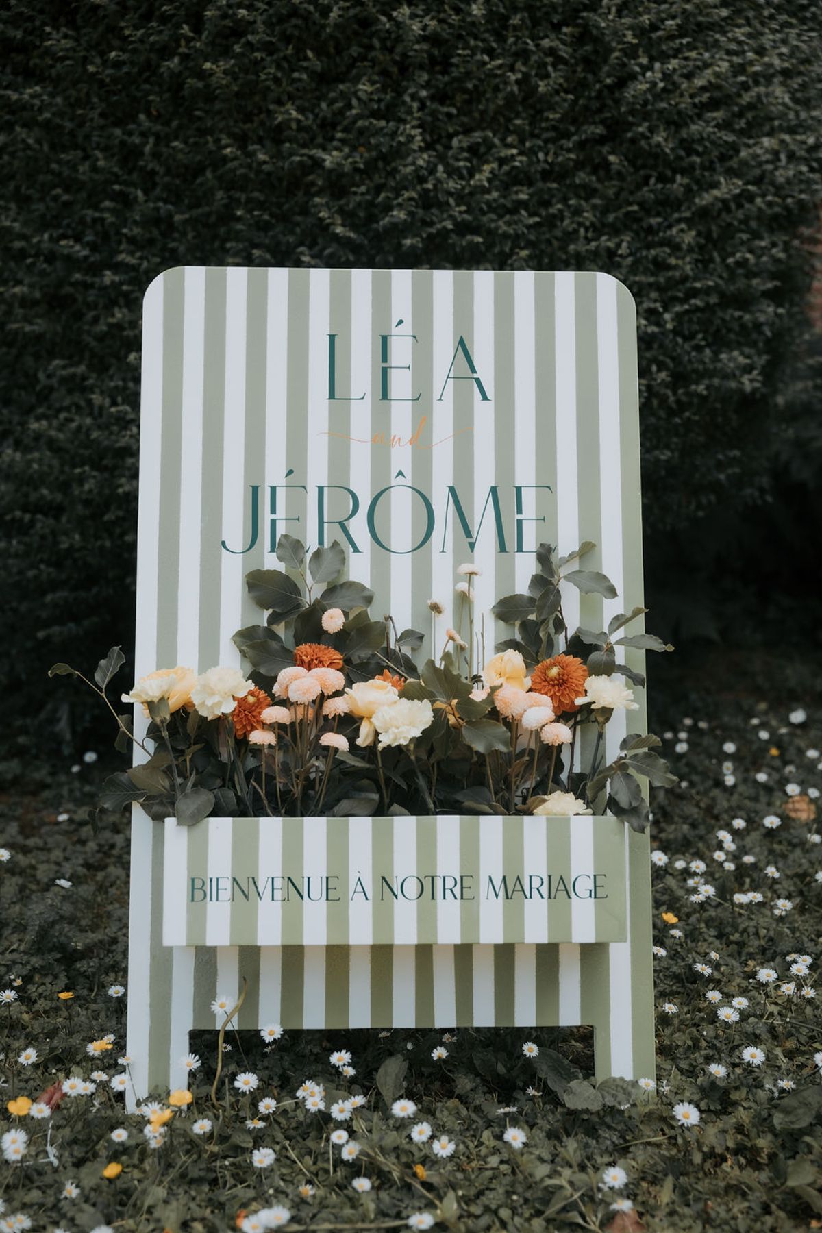 A striped welcome sign reading "Léa & Jérôme, bienvenue à notre mariage" with flowers at its base
