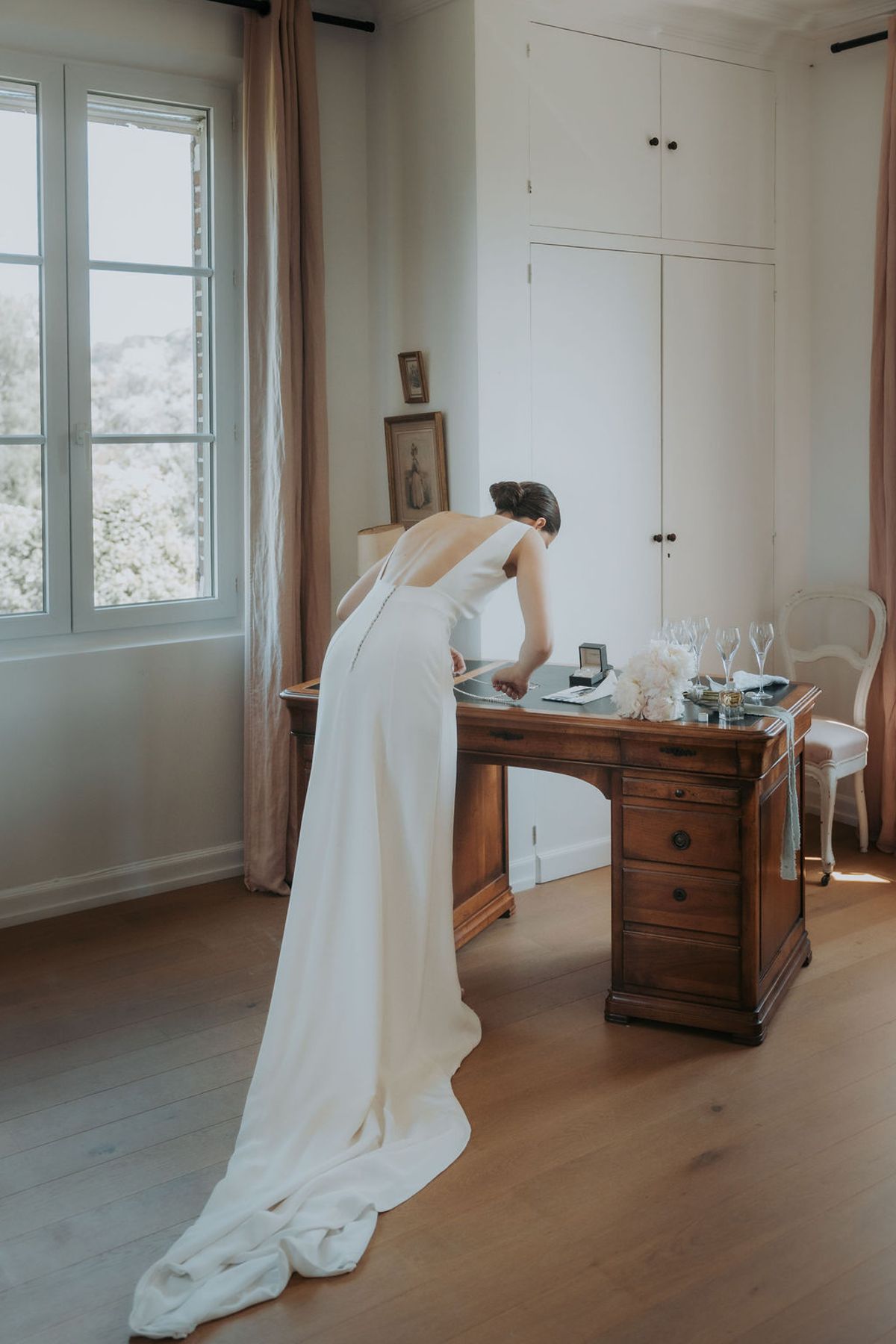 The bride leaning over a wooden desk in a bright bedroom, her train spread across the floor