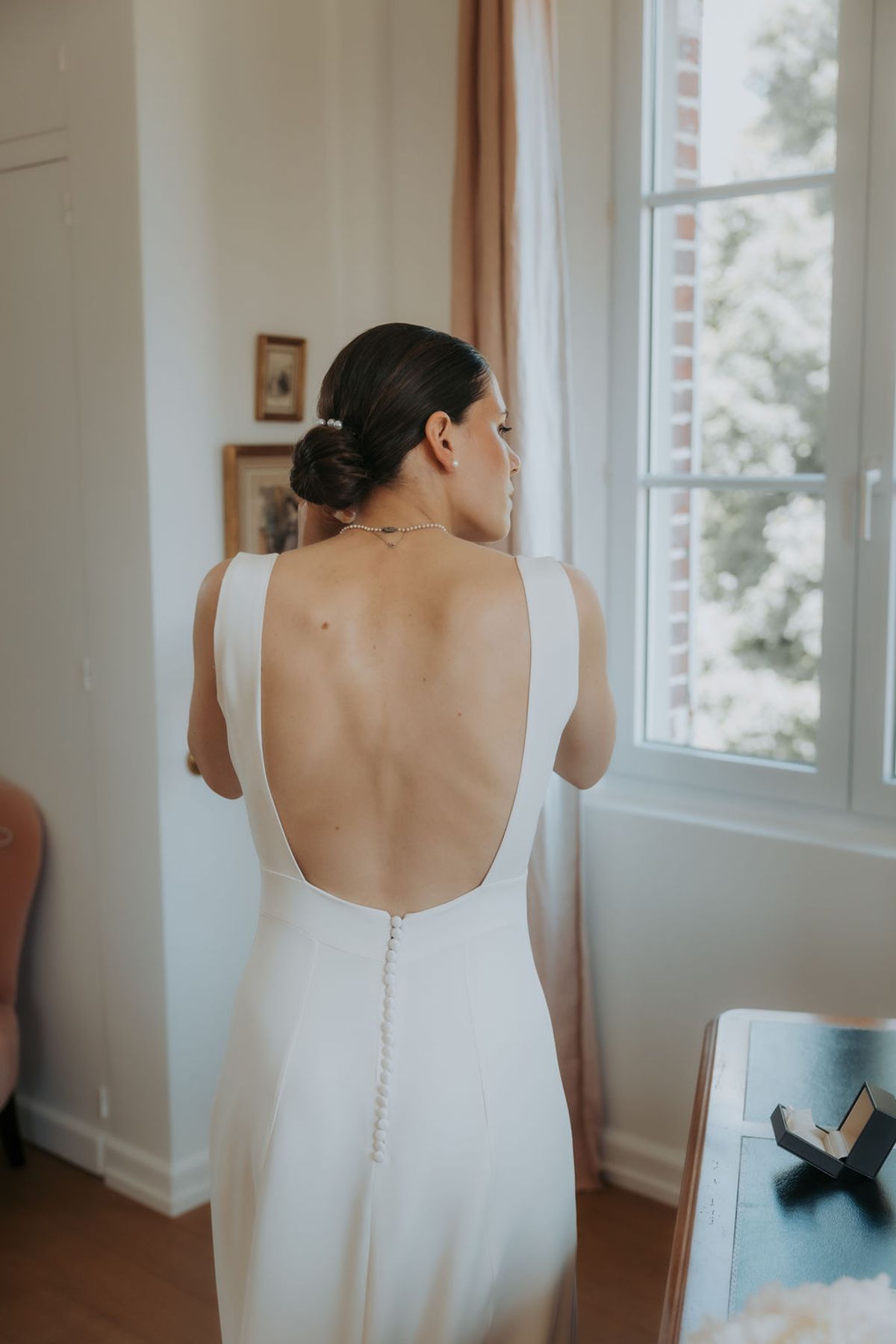 The open back of the bride's dress as she stands by a window