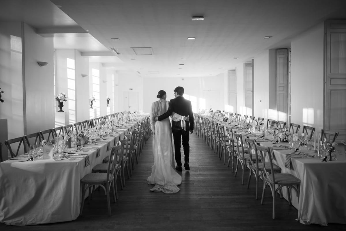 The bride and groom walk between long tables in a bright reception hall, in black and white