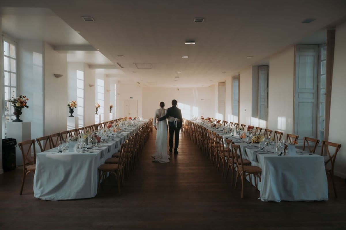 The bride and groom between the long tables set for dinner in the reception hall