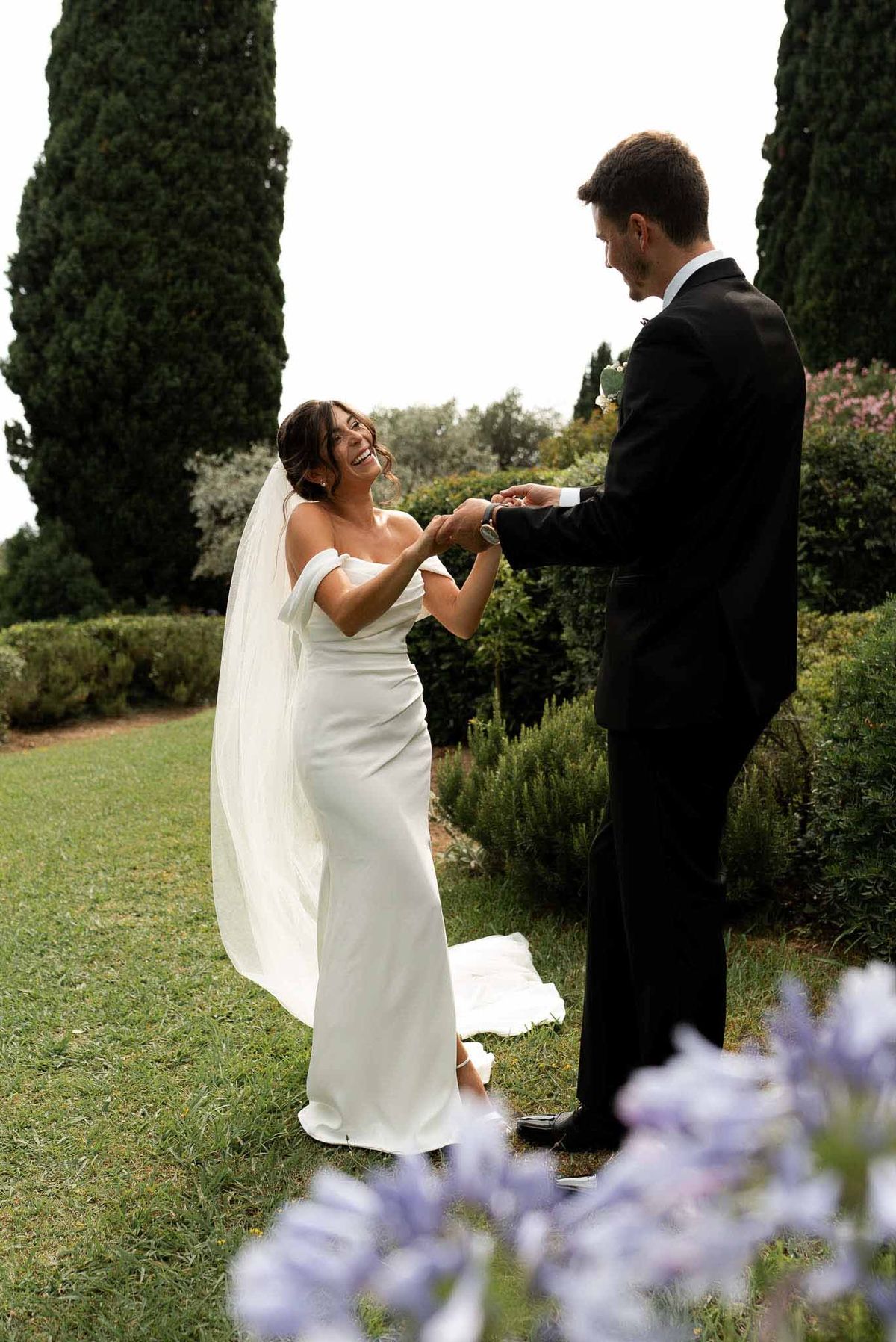 Bride laughing while holding groom's hands in a garden, framed by lavender flowers with cypress trees behind