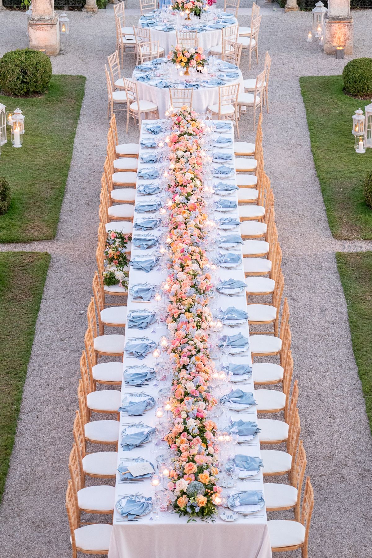 Long banquet table with coral and blush rose runner, dusty blue napkins, and votive candles in formal French garden