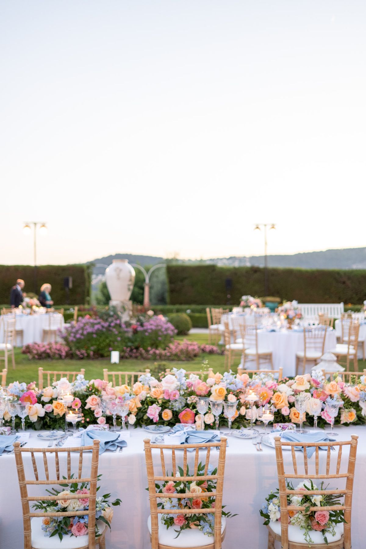 Outdoor reception with long head table and round guest tables on manicured lawn with peach and coral floral runners