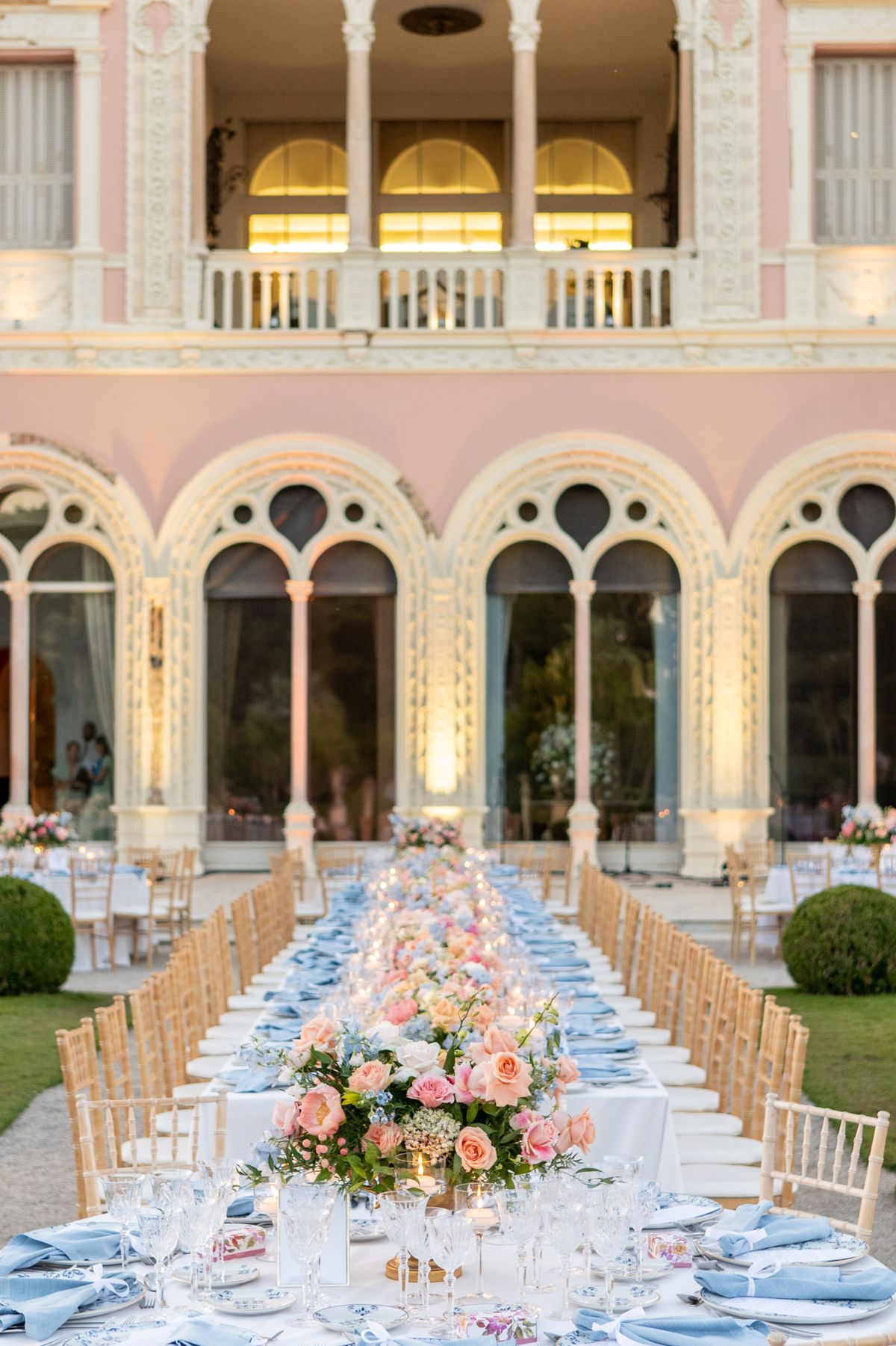 Long feasting table with powder blue napkins and peach rose runner before pink neoclassical villa facade
