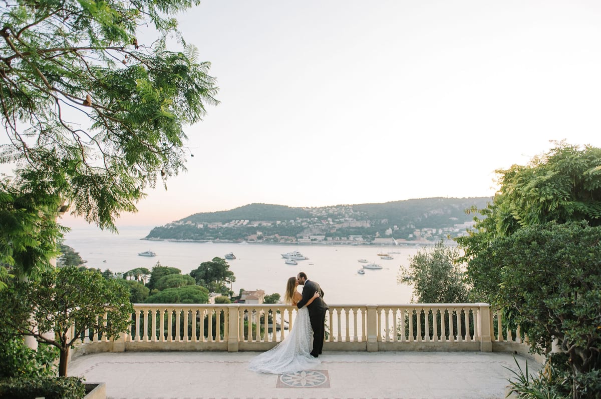 Bride and groom kiss on stone terrace with white balustrade overlooking French Riviera bay with yachts at golden hour