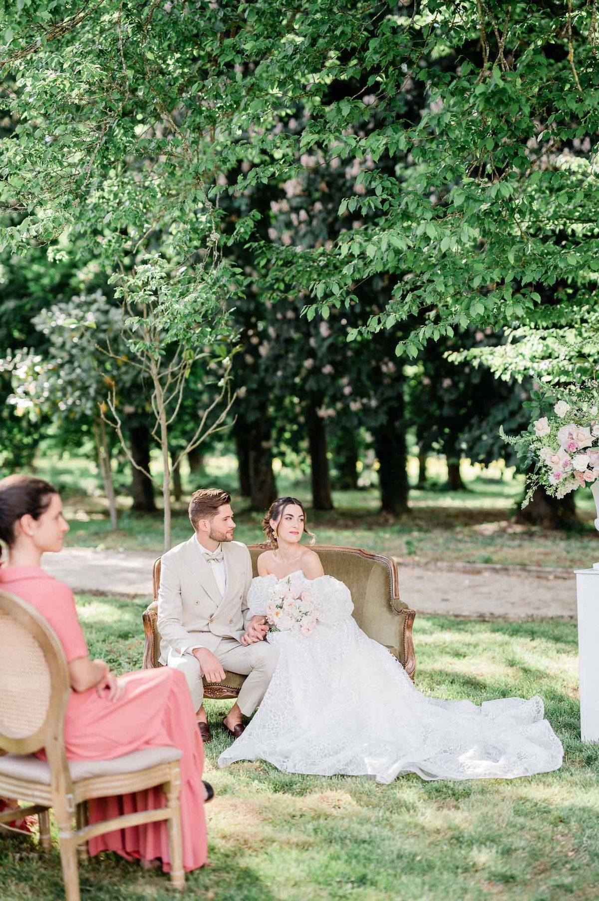 Bride and groom seated together on garden settee with bridesmaid at outdoor wedding reception