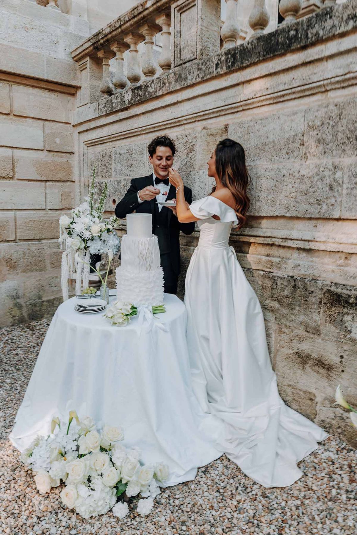 Groom feeding cake to bride beside four-tier white ruffle wedding cake on chateau terrace