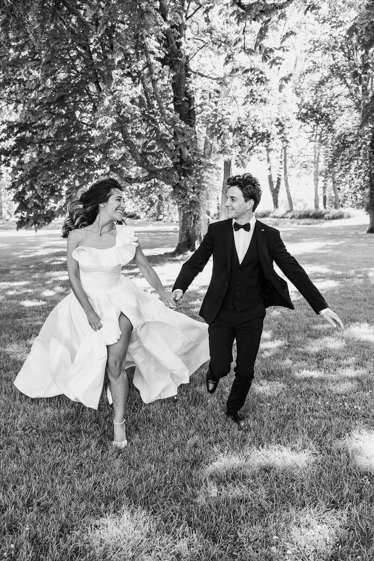 Black and white portrait of bride and groom running hand-in-hand across tree-lined park grounds