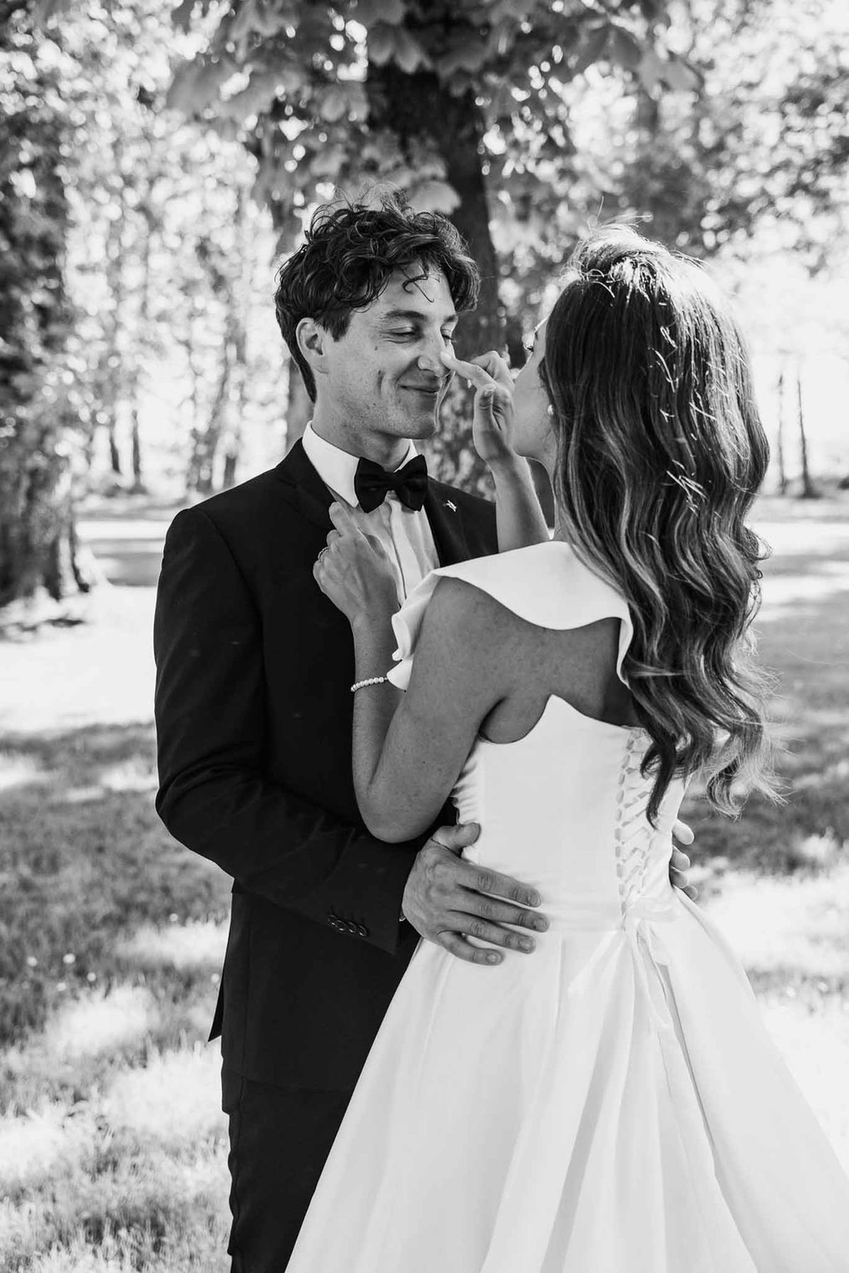 Bride playfully touching groom's nose in backlit wooded park setting in black and white