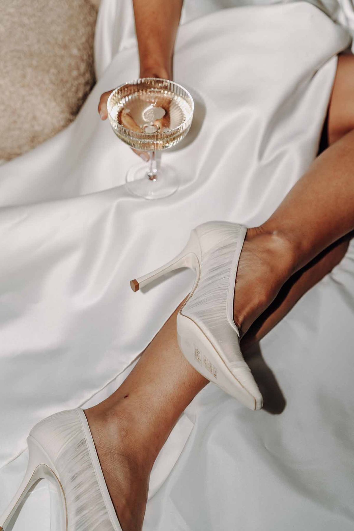 Close-up of bride's ivory satin stiletto heels and champagne coupe with satin dress fabric