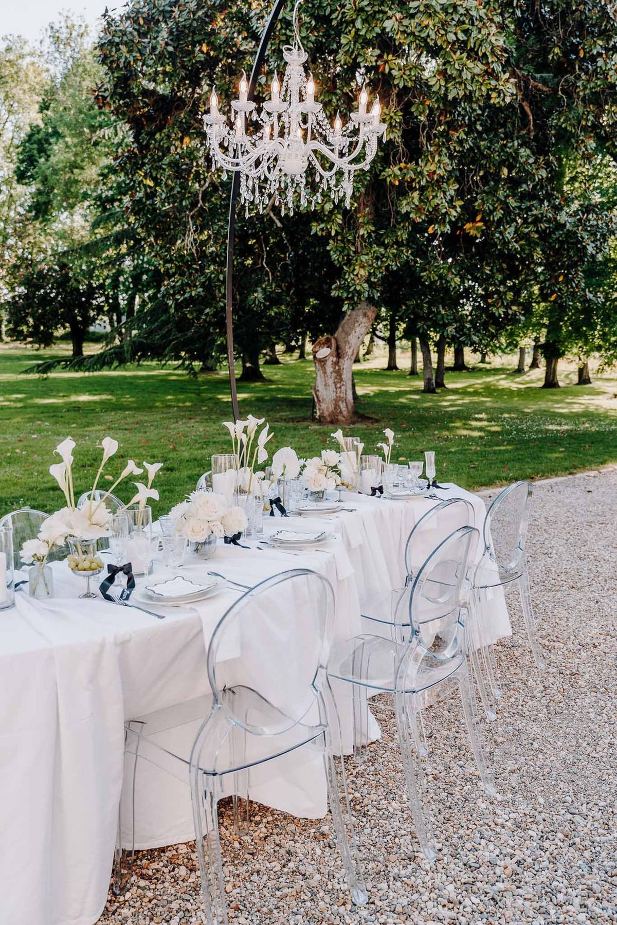 Long table with ghost chairs, white calla lily vases, and suspended crystal chandelier on gravel garden path