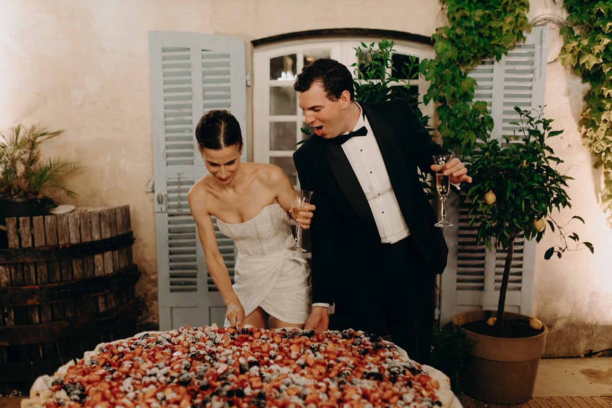 Bride and groom cutting berry-topped tart at night in Provencal courtyard with blue shutters