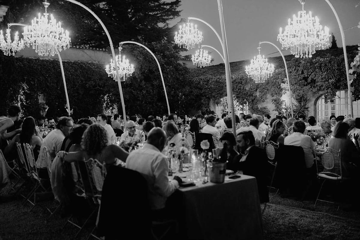 Black and white photo of outdoor chateau reception with guests at round tables under suspended crystal chandeliers