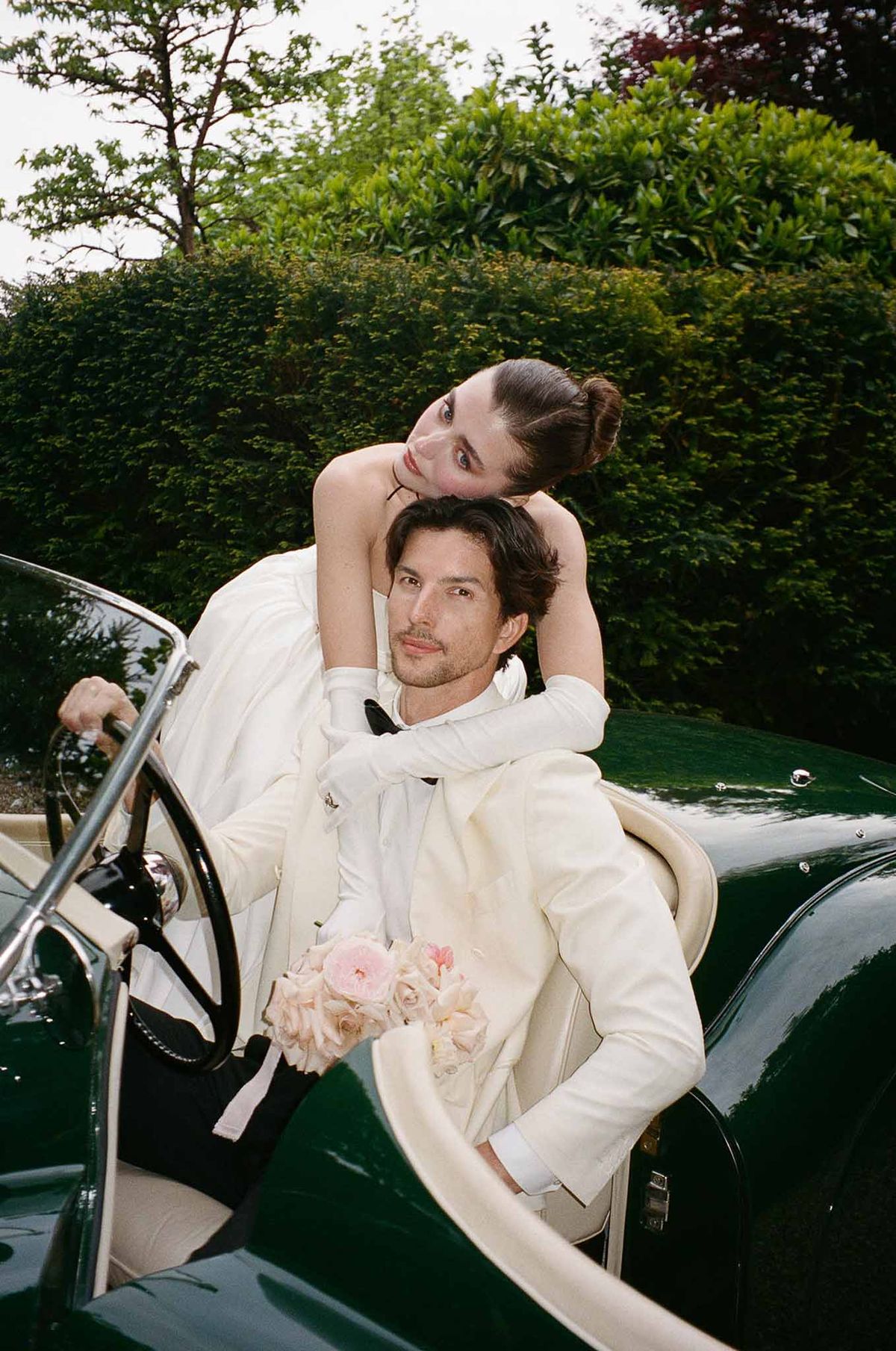 Bride in satin gloves with blush rose bouquet leans over groom in ivory suit in vintage green convertible