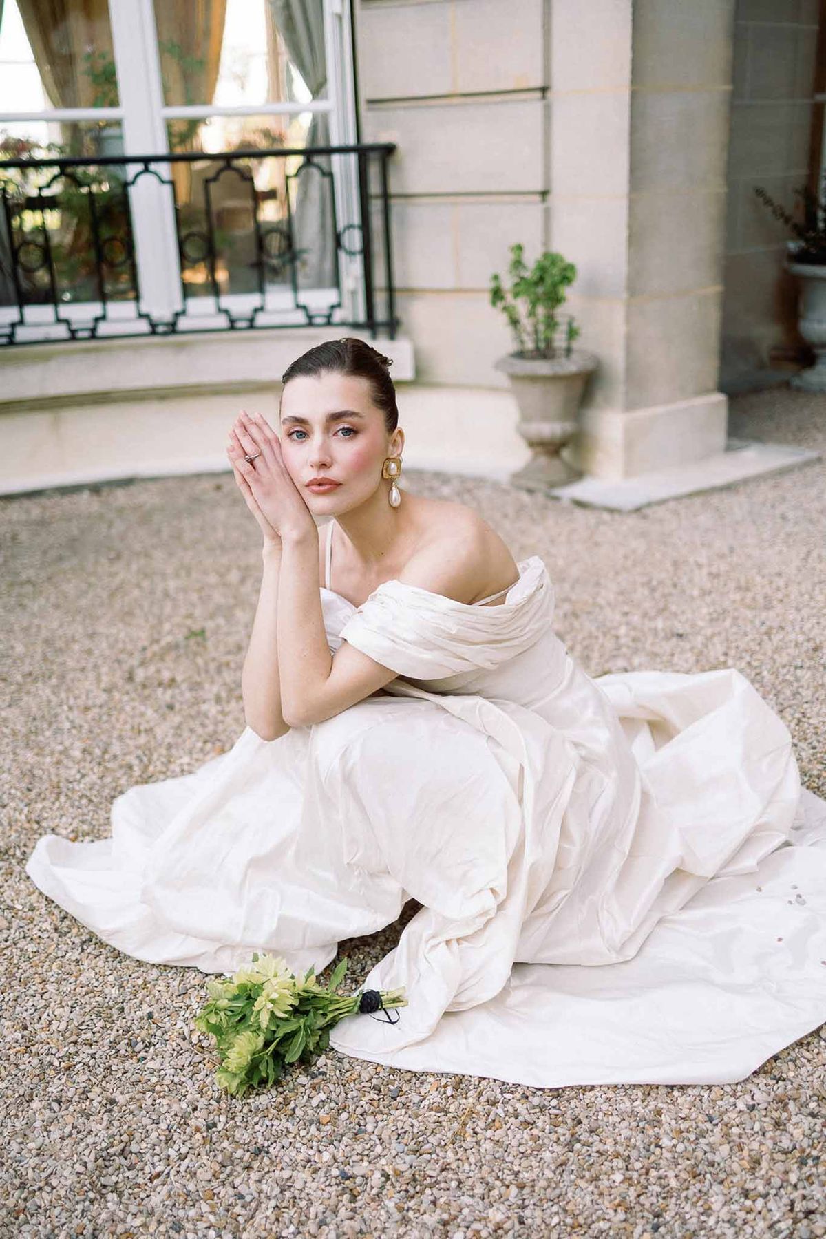 Bride in off-shoulder ballgown with gold pearl earrings seated on gravel before chateau balcony