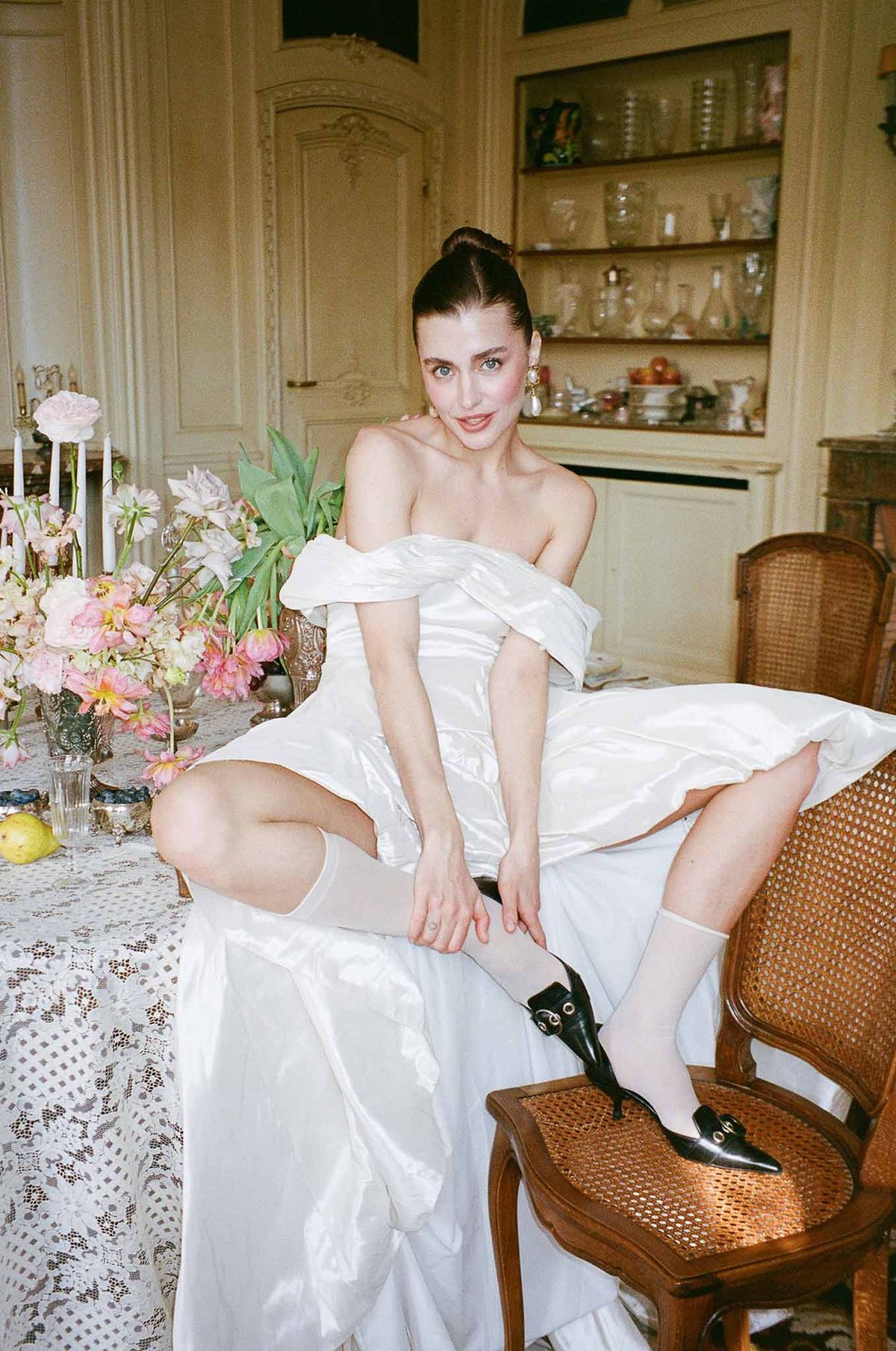 Bride in off-the-shoulder satin gown seated at a lace-covered table with pink peony arrangements