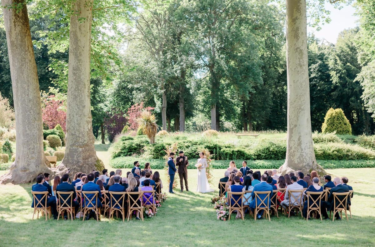 Outdoor wedding ceremony under plane trees with 60 guests, cross-back chairs, and a pink floral arch