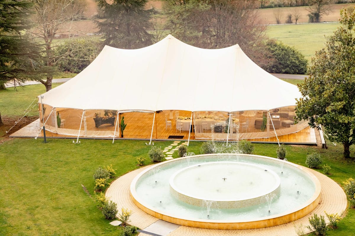 Aerial view of stretch tent reception setup on wooden deck behind a tiered fountain on estate grounds