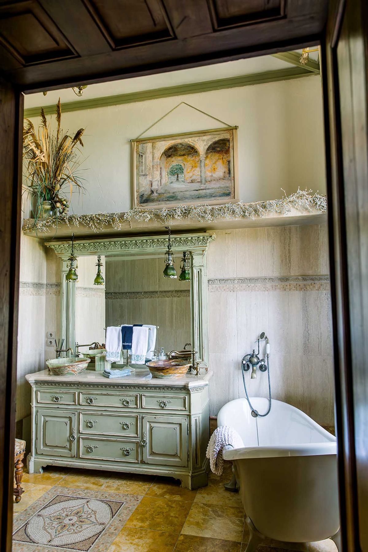 French country bathroom with sage green carved vanity, clawfoot tub, travertine walls, and mosaic floor medallion