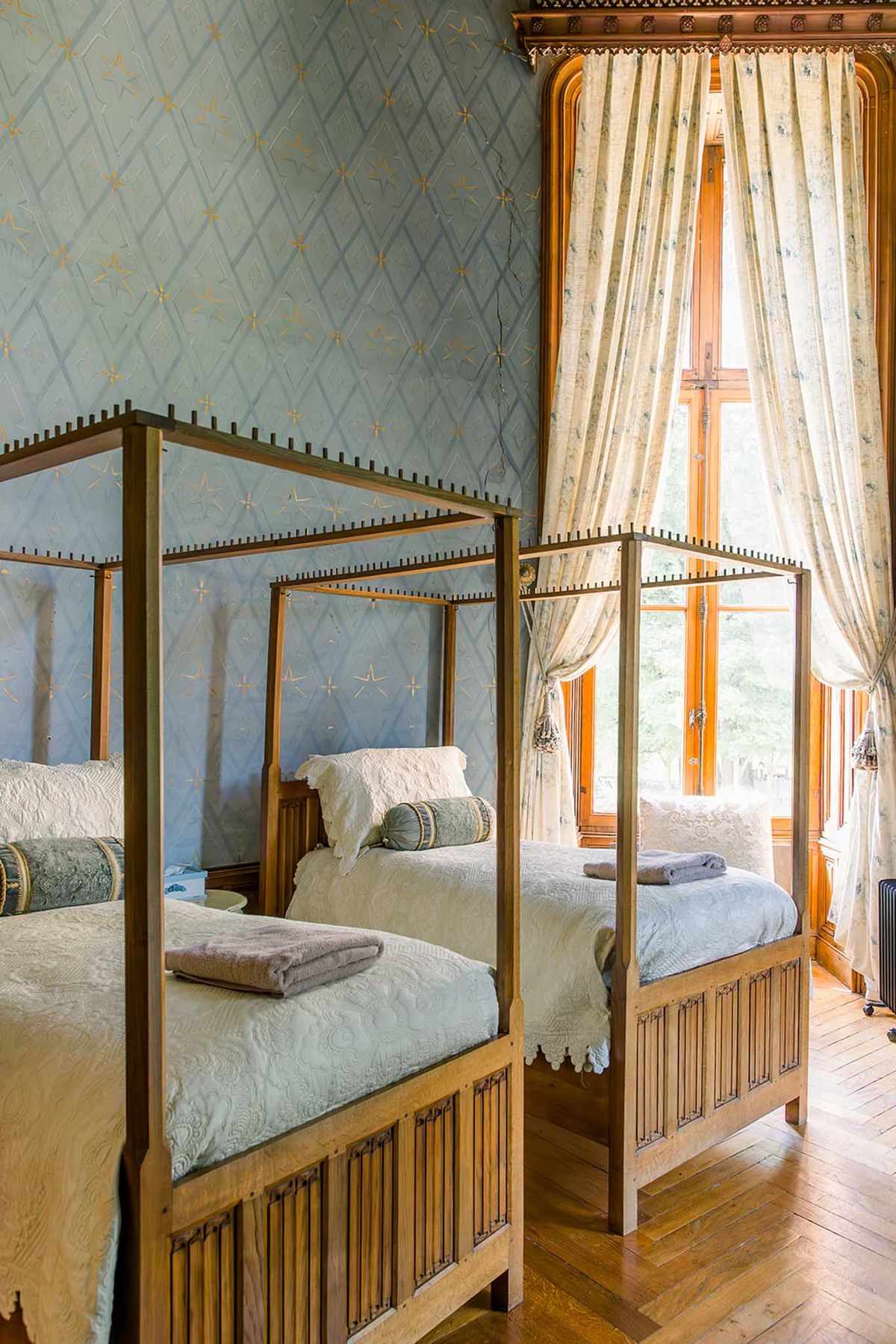 Chateau bedroom with four-poster beds, blue-grey diamond wallpaper, and cream floral curtains