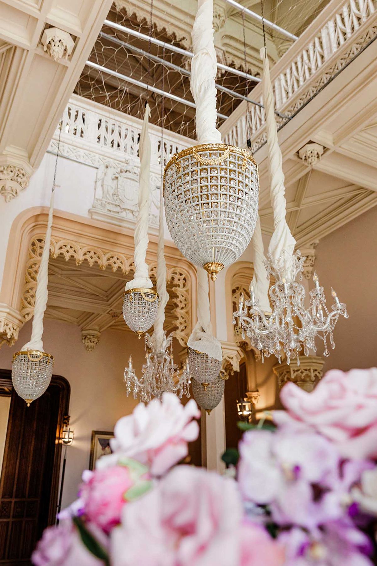 Crystal and gold chandeliers hanging from ornate Gothic-style ceiling with pink floral arrangements below