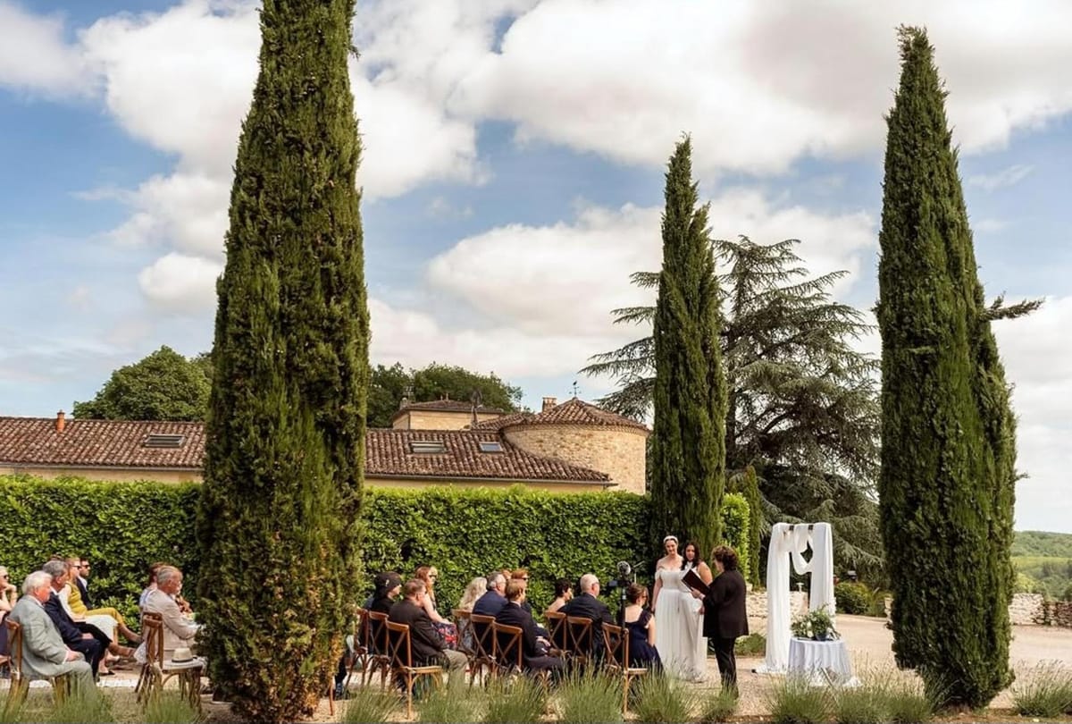 Outdoor wedding ceremony at French chateau with guests seated on wooden chairs facing couple at white-draped altar