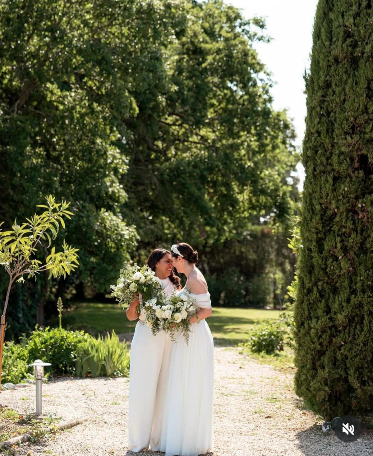 Two brides with white rose and eucalyptus bouquets sharing a kiss on cypress-lined garden path