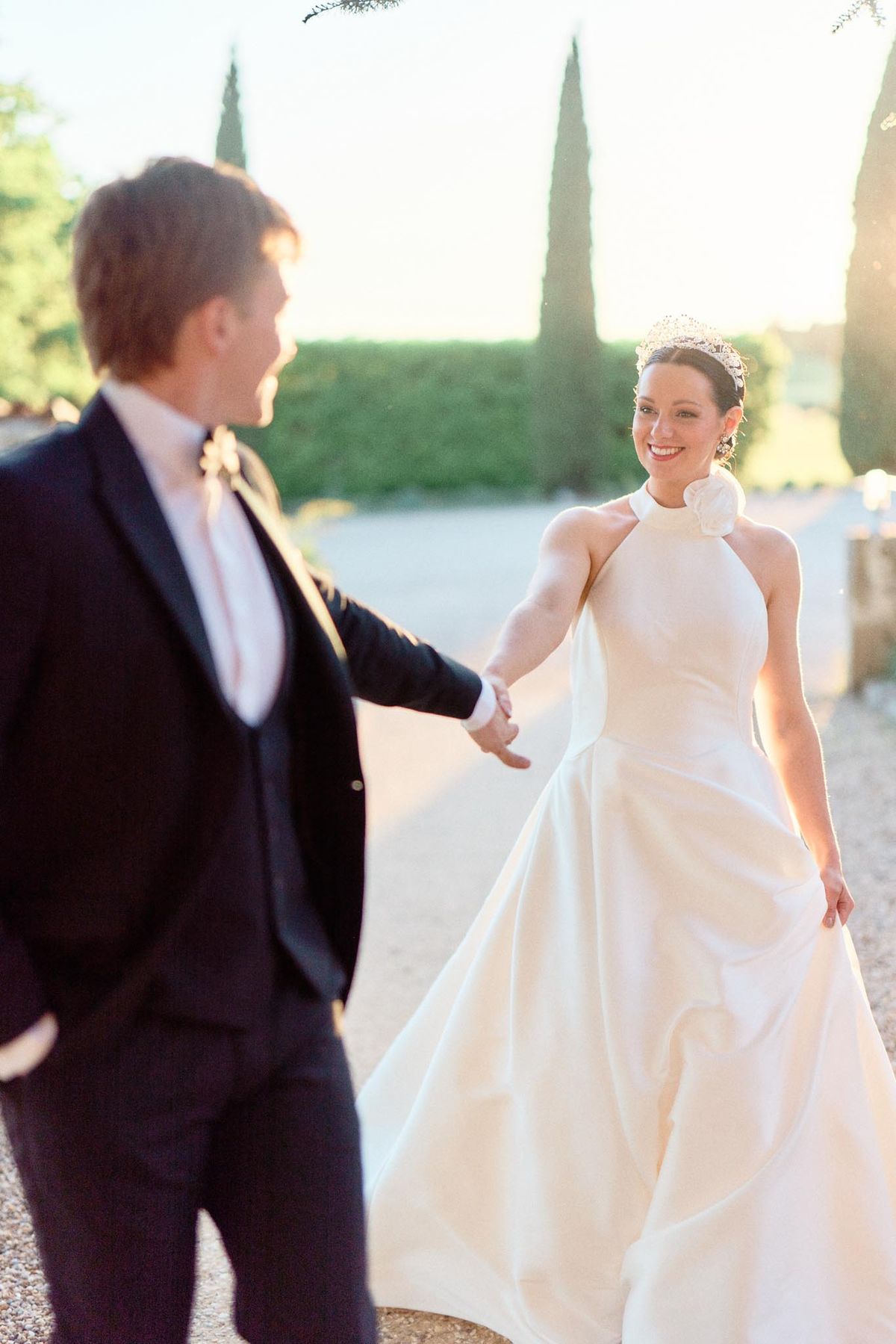 Bride in ivory halter-neck gown and groom in navy suit holding hands on gravel path at golden hour