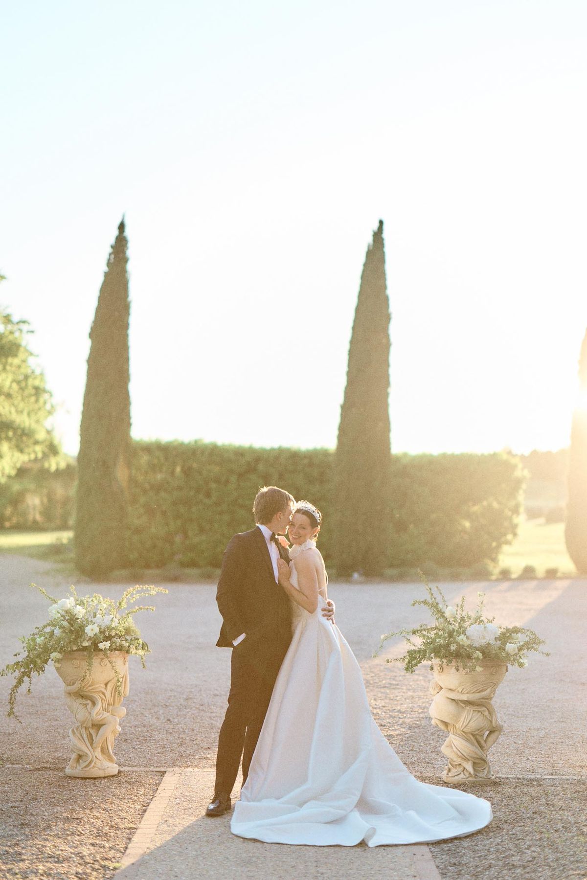 Couple kissing at golden hour between stone urn floral arrangements on formal chateau axis pathway