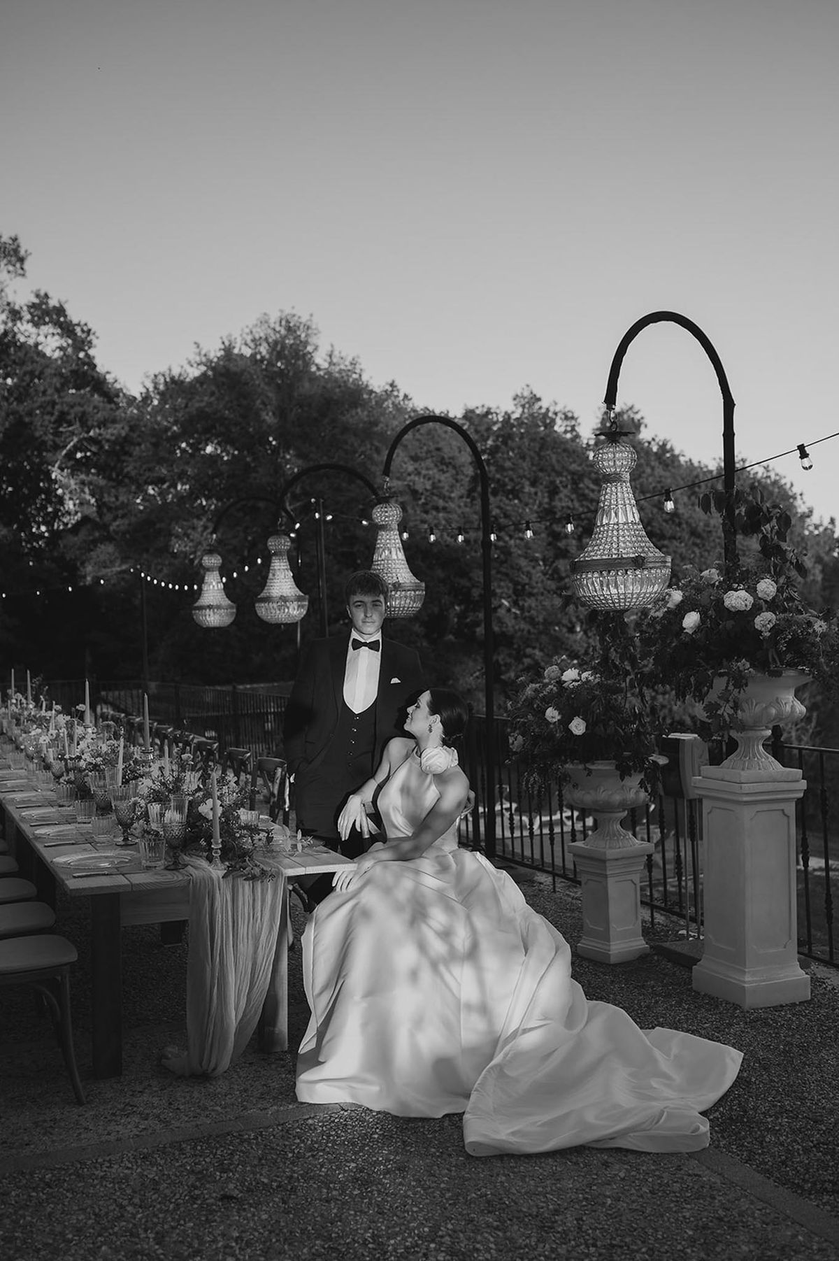 Bride in ballgown and gloves sits as groom stands beside candlelit banquet table with chandeliers in B&W